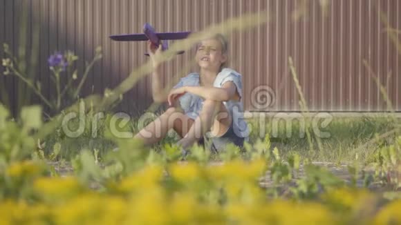 一个漂亮可爱的小女孩坐在篱笆下的草地上发射小飞机的肖像孩子花钱视频的预览图