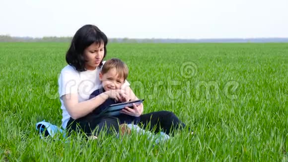 幸福家庭母子坐在绿草上妈妈和男孩用平板画技术应用视频的预览图