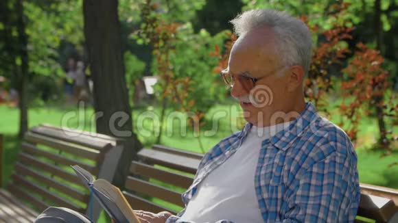 一位老人坐在长凳上在公园里读一本有趣的书视频的预览图