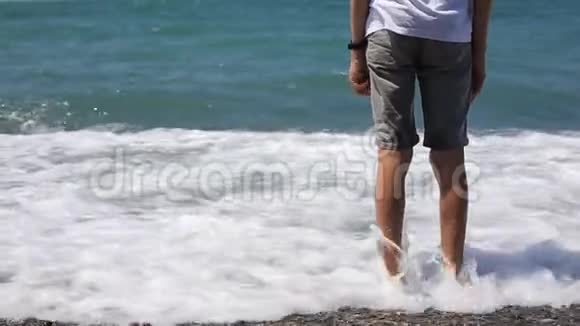 男孩脚在海里一个十几岁的少年站在海滩上的鹅卵石海滩上自然假期视频的预览图