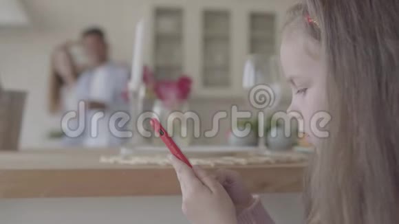小女孩玩手机背景是她的父母在大厨房里视频的预览图