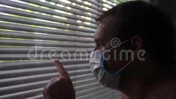 戴着防护口罩的悲伤青年看着窗外的百叶窗视频的预览图