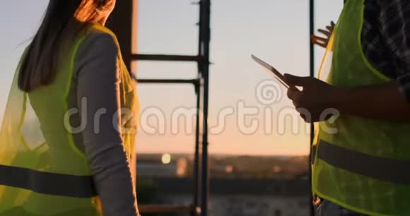 建筑工人拿着一块石碑的男人和一个戴着白色头盔的女人在夕阳下站在大楼的屋顶上握手视频的预览图
