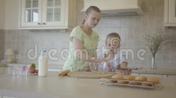 母亲在现代舒适的厨房里帮助和教她可爱的小女儿做蛋糕或煎饼幸福家庭视频的预览图