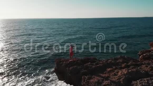 在岩石海岸穿红色裙子的漂亮女孩穿红色衣服的女士站在悬崖上女孩看着海洋日落鸟瞰图视频的预览图
