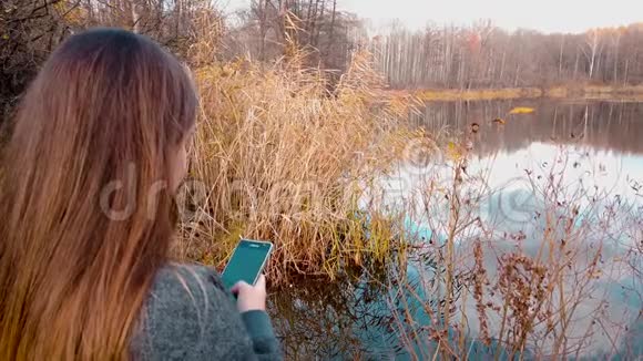 一个女孩在美丽的森林湖畔在智能手机上的社交网络聊天中写道视频的预览图