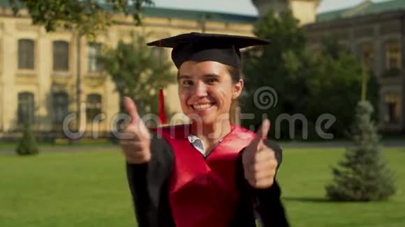 戴着黑色斗篷和帽子的漂亮女孩站在大学大楼前看镜头的肖像视频的预览图
