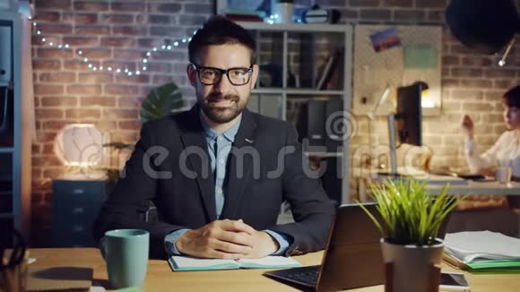 快乐的商人戴着眼镜晚上坐在办公桌前微笑视频的预览图