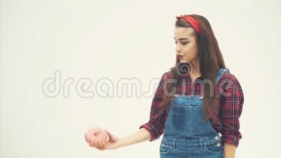 幸福的年轻女士站着舔着嘴唇举起手里拿着一个非常美味的粉红色甜甜圈视频的预览图