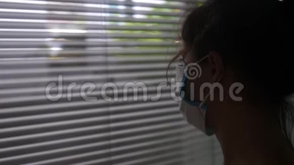 在家的年轻女子戴着医疗面罩看着窗外由于冠状病毒的传播而被隔离流行病视频的预览图