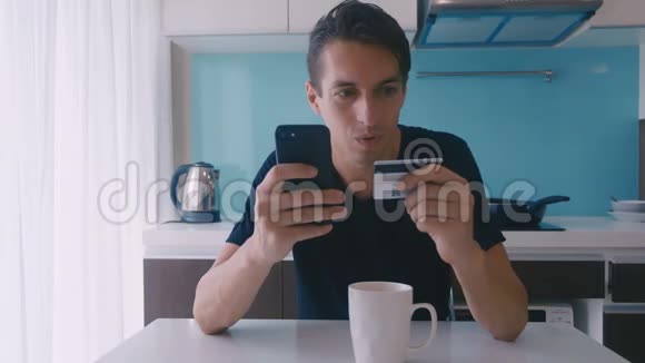 年轻的帅哥坐在桌子旁用智能手机在厨房的家里用信用卡进行网上购物室内视频的预览图