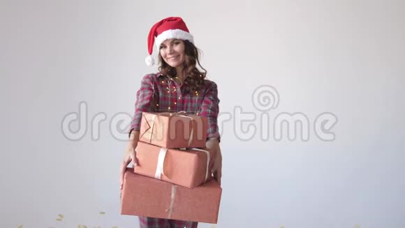 穿着睡衣和圣诞帽的年轻女子把圣诞礼物放在白色背景上女孩带着礼物跳舞视频的预览图