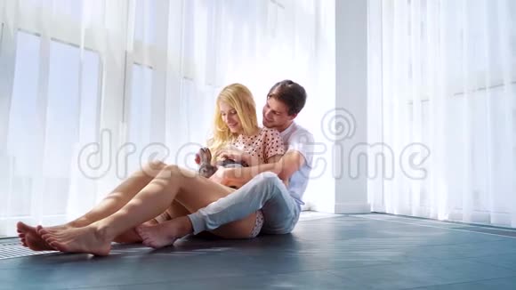 穿着睡衣和家兔坐在地板上的可爱情侣的全景镜头视频的预览图