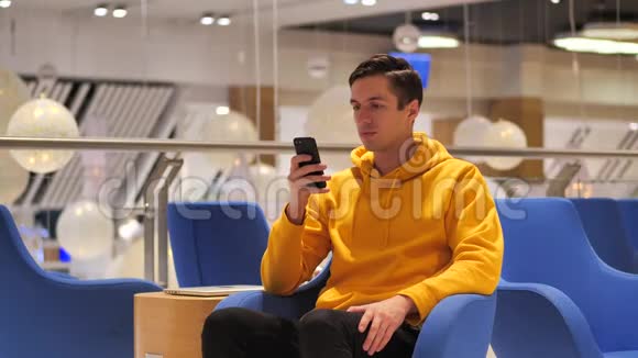 穿黄色连帽衫的年轻男性在舒适的咖啡馆里使用智能手机沟通商务旅行约会的概念视频的预览图