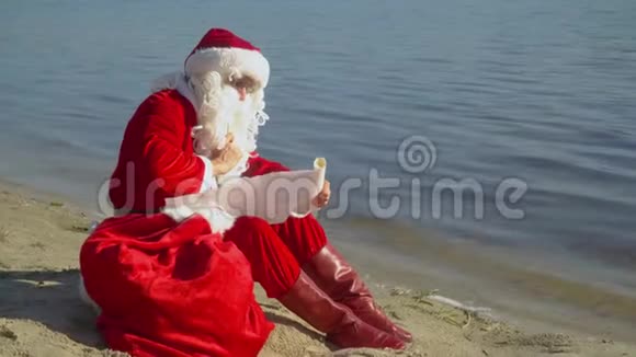 圣诞老人坐在海边的沙滩上拿着一袋礼物在名单上写了一些东西视频的预览图