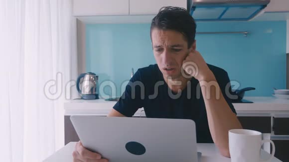 沮丧的年轻人在家里厨房里用笔记本电脑工作一个人受苦头痛视频的预览图