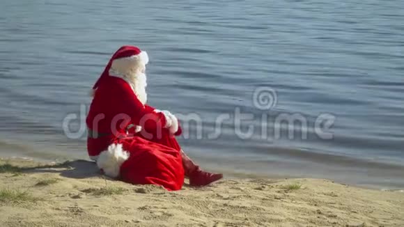 圣诞老人坐在河边或湖边拿着一袋礼物圣诞老人望着远处的水视频的预览图