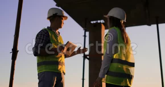 建筑工人拿着一块石碑的男人和一个戴着白色头盔的女人在夕阳下站在大楼的屋顶上握手视频的预览图