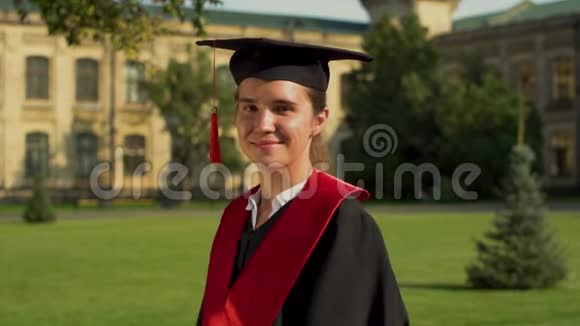 戴着黑色斗篷和帽子的漂亮女孩站在大学大楼前看镜头的肖像青年视频的预览图