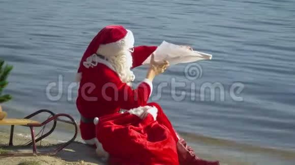 圣诞老人坐在湖边的沙滩上带着一个礼品袋看着名单视频的预览图