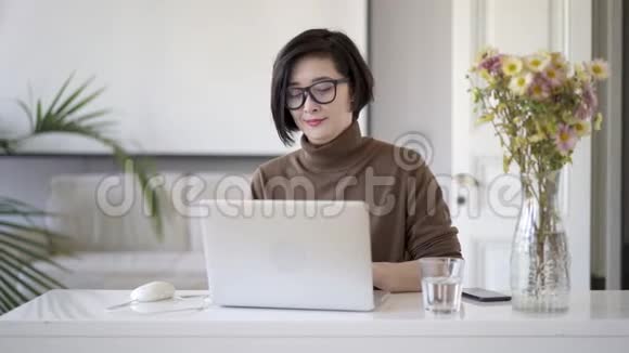 戴眼镜的亚洲女性在白色家庭办公室用笔记本电脑写字视频的预览图