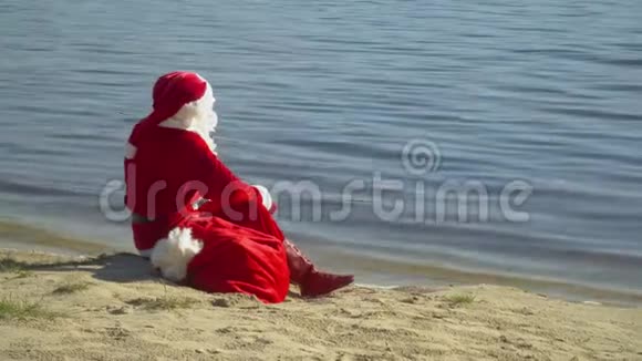圣诞老人坐在河边或湖边拿着一袋礼物圣诞老人看着远处的水圣诞老人视频的预览图