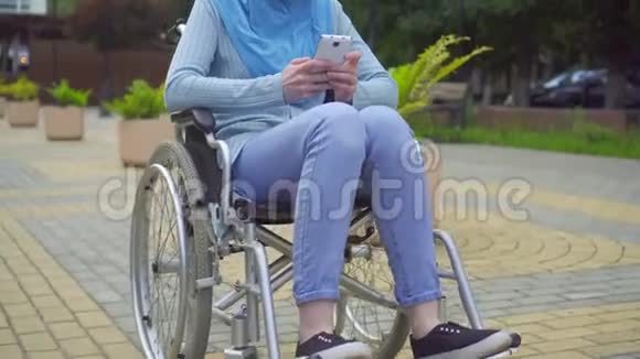 肖像年轻迷人的穆斯林妇女残疾的传统围巾坐在轮椅上使用智能手机视频的预览图