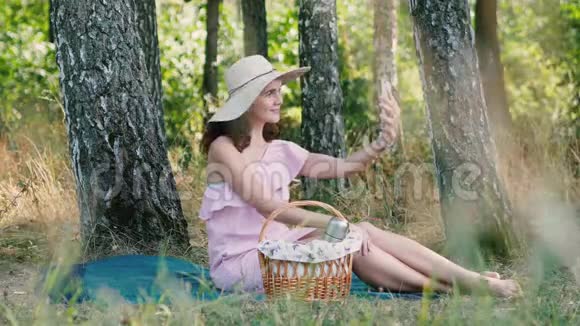 野餐时间年轻漂亮的女孩戴着圆帽子坐在格子上摆出自拍的姿势公园的户外活动视频的预览图