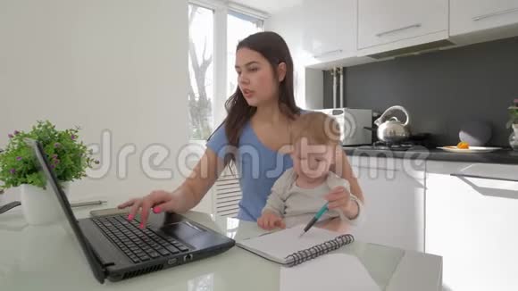 妈妈和爱哭的孩子结合了父母的养育和在厨房的桌子旁工作的笔记本电脑视频的预览图