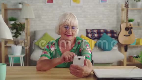 体贴的老妇人在客厅的桌子旁使用智能手机视频的预览图