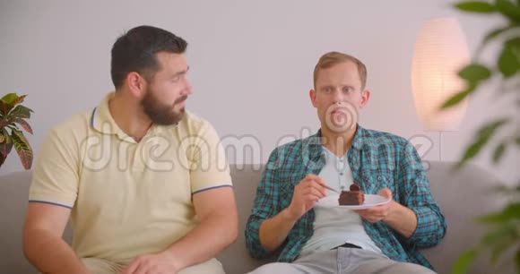 两个高加索男人坐在沙发上看电视的特写镜头伤心的男人吃巧克力蛋糕视频的预览图