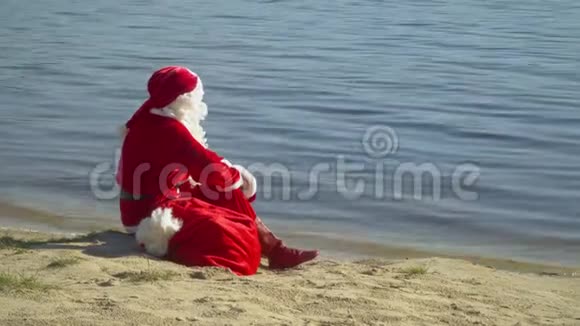 圣诞老人坐在河边或湖边拿着一袋礼物圣诞老人望着远处的水视频的预览图