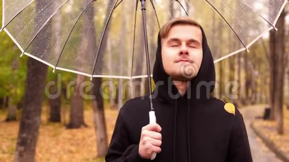 一位年轻英俊的男子撑着伞在雨天沿着秋天公园散步的肖像视频的预览图