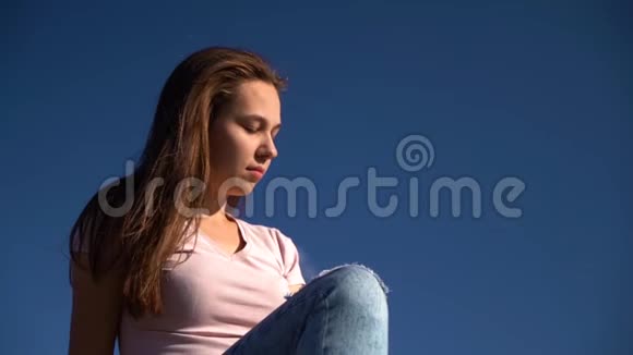 漂亮的女孩或年轻的女人穿着粉红色的衬衫和蓝色撕裂的牛仔裤坐在田野中间的木头建筑上视频的预览图