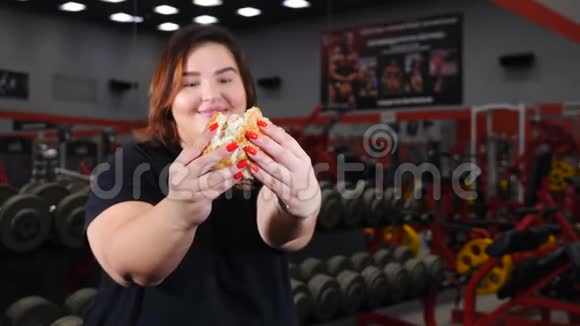 肥胖妇女在健身房吃汉堡包微笑和看相机的肖像食物诱惑超重概念视频的预览图