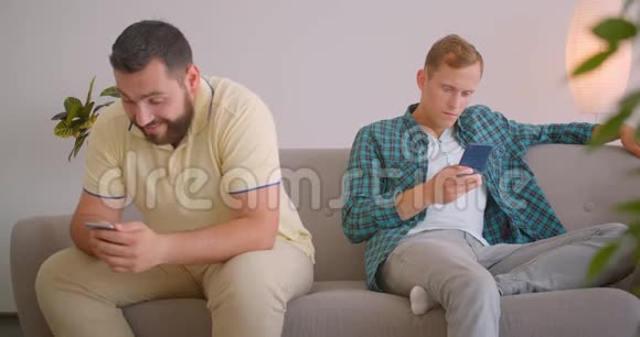 两个白种人使用手机坐在舒适的家庭室内沙发上的特写镜头朋友分享有趣视频的预览图