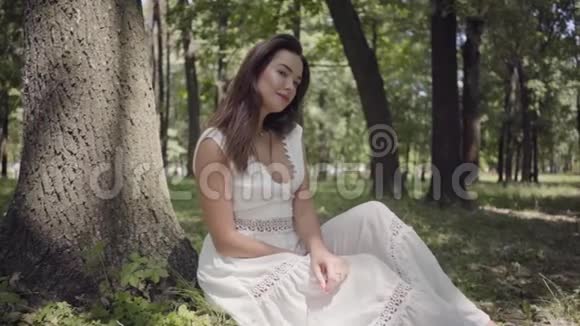 一位美丽的少女一头深色长发穿着白色的夏装坐在树下视频的预览图