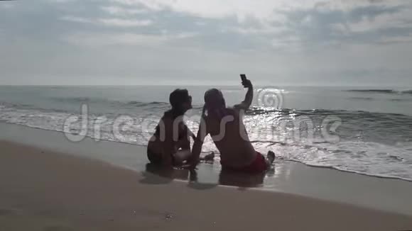 一对夫妇坐在海滩上在日出时用智能手机拍照视频的预览图