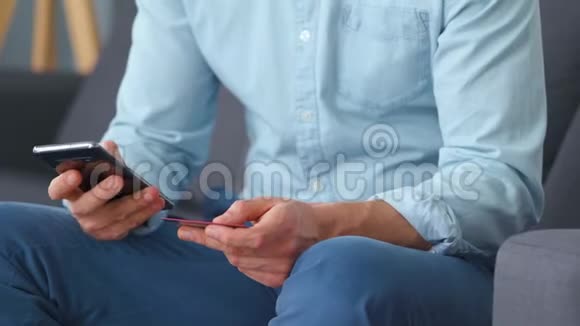 正式穿着的男人坐在沙发上把信用卡号码输入智能手机在线支付网上购物视频的预览图