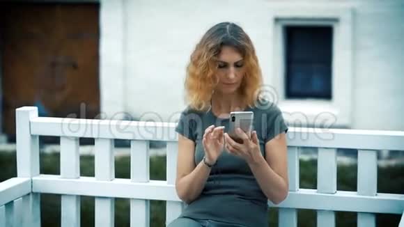 一个身材苗条悲伤金发卷曲的中年妇女晚上坐在一张白色的长凳上看着电话视频的预览图