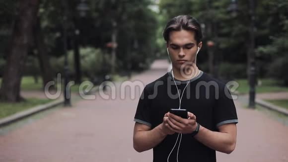 戴着耳机和智能手机的帅哥用它站在公园慢动作概念健康视频的预览图
