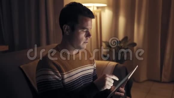 一位严肃的年轻人坐在沙发上深夜忙于制作数码平板电脑视频的预览图