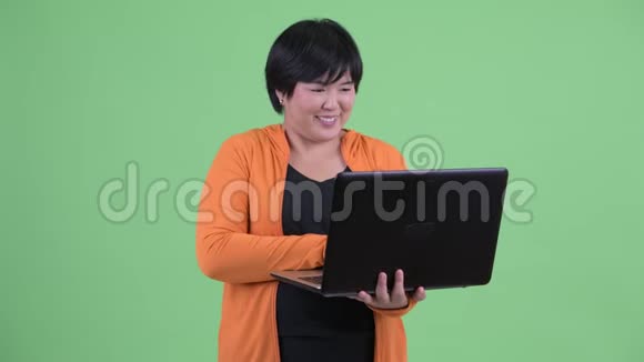 快乐年轻体重超重的亚洲女人一边想着一边用笔记本电脑准备健身房视频的预览图