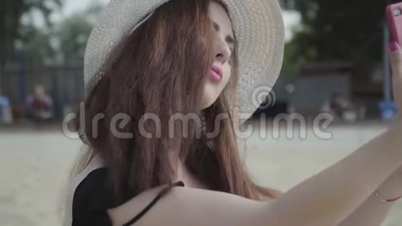 可爱的年轻女人在夏天的白色帽子自拍她的手机休息在海滩上夏季休闲概念视频的预览图