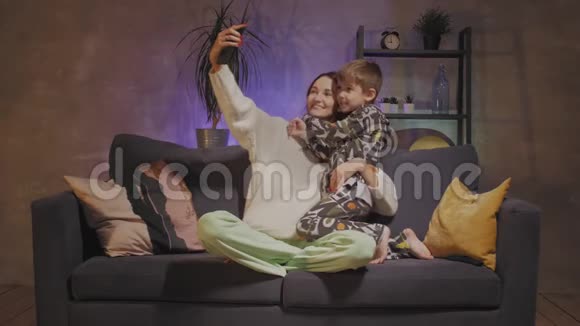 年轻的母亲和儿子坐在舒适的房间里的沙发上自拍视频的预览图