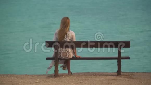 孤独的女孩穿着长长的白色连衣裙坐在长凳上回到海上或海洋背景上的相机金发女人的休闲视频的预览图