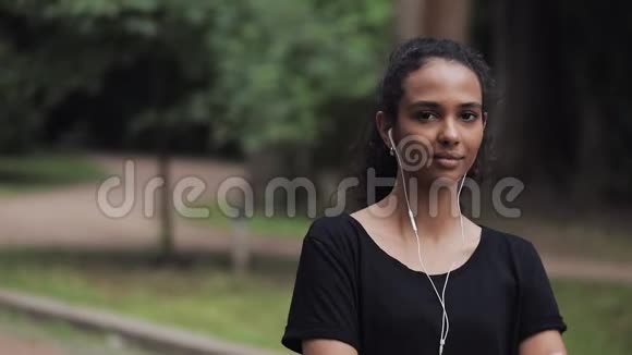 戴着耳机站在公园里的漂亮年轻音乐女孩的肖像转过头微笑健康的理念视频的预览图