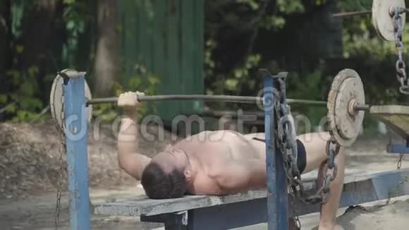 强壮的人举起杠铃躺在公园的长凳上锻炼肱二头肌保持身体的形状健身视频的预览图