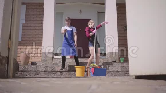 美丽的年轻女子在房子的门廊上跳舞唱歌而她的丈夫喷洒洗涤剂清洁视频的预览图