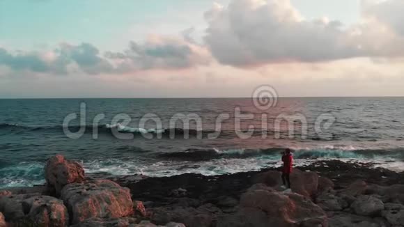一位年轻的白种人女摄影师正站在岩石海岸的大石头上拍摄美丽的粉红色照片视频的预览图